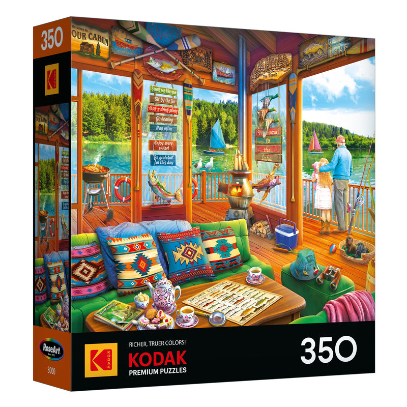 The ROSEART-KODAK PREMIUM-THE LAKE HOUSE 350 piece jigsaw puzzle features a vibrant lakeside cabin scene with people on a dock and cozy interior decor.