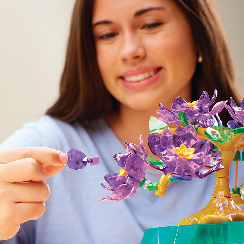The patient artisan assembles a Crystal Gem Creations Blossoming Bonsai, adorned with purple flowers. Each delicate piece added enhances the miniature into an enchanting work of art.