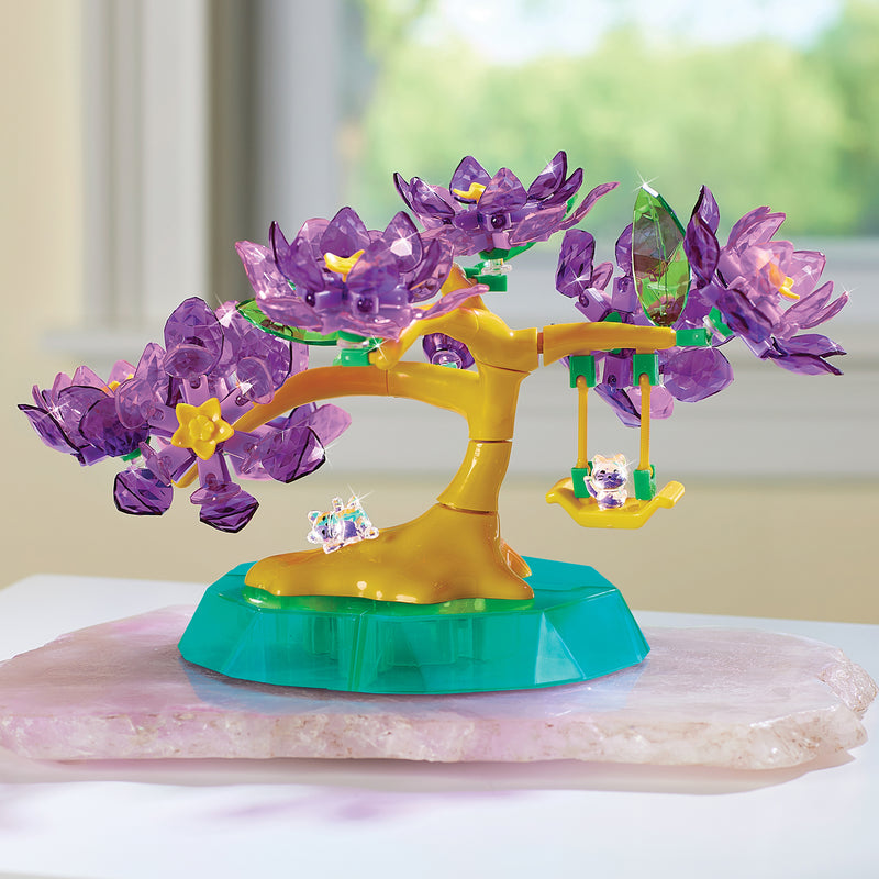 The Crystal Gem Creations Blossoming Bonsai is a plastic toy tree with purple translucent leaves and a yellow trunk on a green base. It includes a swing and small flower, elegantly resting on a pale pink surface indoors.