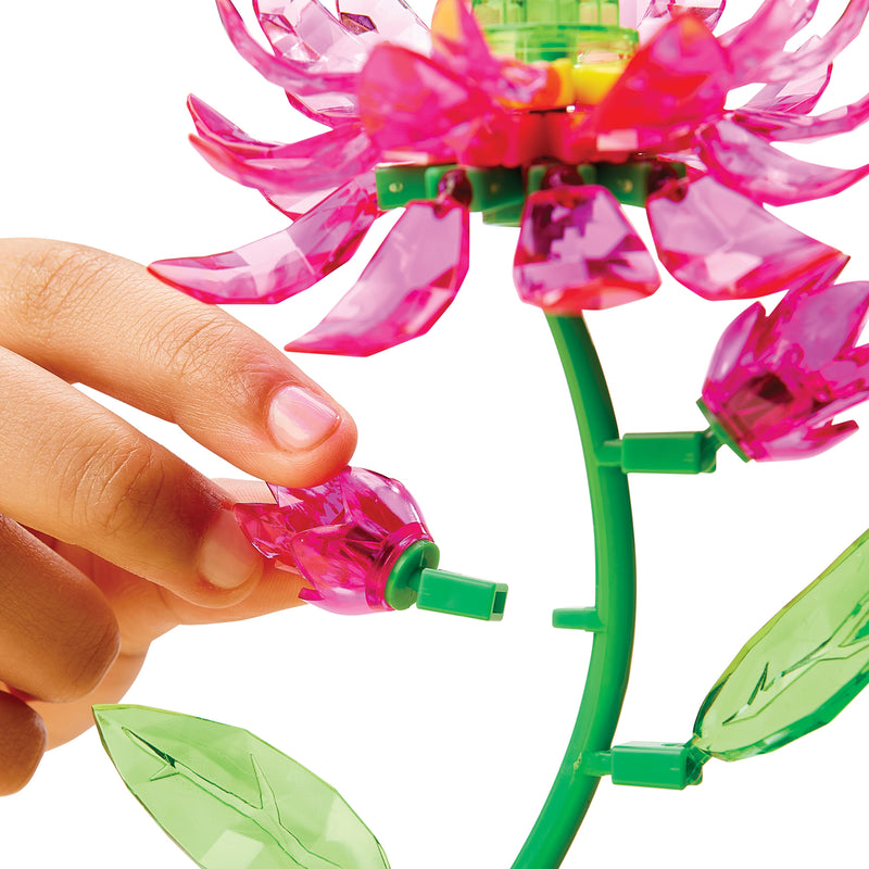 A hand assembles a translucent pink plastic flower using interlocking blocks, resembling the Crystal Gem Creations Blooming Lotus with its vibrant green stem and leaves.