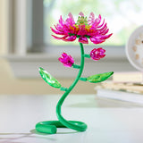 A Crystal Gem Creations Blooming Lotus, featuring a decorative pink and green plastic flower with a flexible stem, sits gracefully on a white surface near a window.