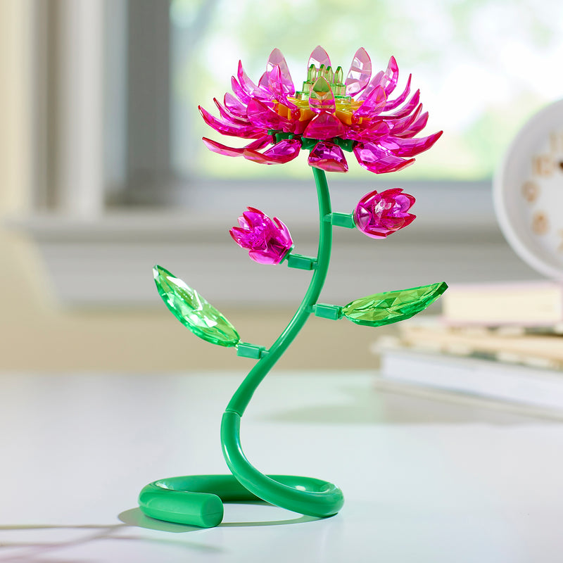 A Crystal Gem Creations Blooming Lotus, featuring a decorative pink and green plastic flower with a flexible stem, sits gracefully on a white surface near a window.