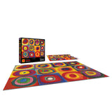 A finished ROSEART - KODAK Masters Color Study, Squares with Concentric Circles 2000-piece jigsaw puzzle lies on the floor next to its box, showcasing vibrant multicolored concentric circles reminiscent of a stunning RoseArt creation.