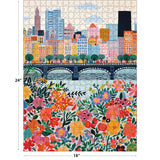 The ROSEART VERTICAL VISIONS CITY FLOWERS 500 piece jigsaw puzzle showcases a vibrant city skyline with tall buildings, a bridge spanning water, and colorful flowers in the foreground.