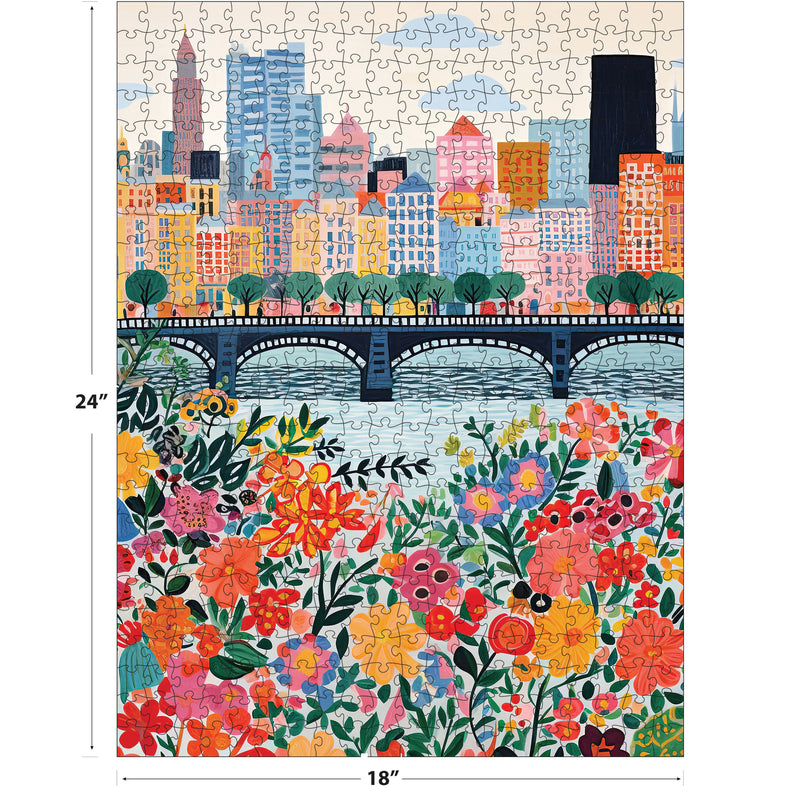 The ROSEART VERTICAL VISIONS CITY FLOWERS 500 piece jigsaw puzzle showcases a vibrant city skyline with tall buildings, a bridge spanning water, and colorful flowers in the foreground.