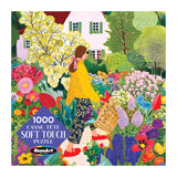 A woman with long brown hair walks through a vibrant garden in front of a white house on the ROSEART Soft Touch Picking Flowers 1000 piece jigsaw puzzle box.