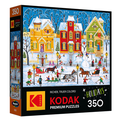 The ROSEART - KODAK PREMIUM "After the Snow Falls" 350-piece jigsaw puzzle features a festive winter village scene with snow, a horse-drawn sleigh, and people playing outside.