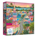 The ROSEART-MILLIE ILLUSTRATES-CHARLESTON-300 PIECE JIGSAW PUZZLE features an illustrated cityscape of Charleston with historic buildings, lush greenery, and a waterfront at sunset.