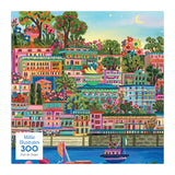 ROSEART-MILLIE ILLUSTRATES-PORT DE SOLLER-300 PIECE JIGSAW PUZZLE features a colorful painting of Port de Soller, a Mediterranean town with vibrant buildings, lush greenery, and boats on the water beneath a blue sky.