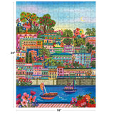 The ROSEART-MILLIE ILLUSTRATES-PORT DE SOLLER 300 Piece Jigsaw Puzzle features a vibrant coastal town with buildings, greenery, flowers, and boats on blue water. Finished size: 24" x 18".