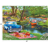 The ROSEART-BIGELOW ILLUSTRATIONS-GOOD TIMES AT THE RIVER 500-piece jigsaw puzzle (18"x24") features a vintage blue car by a picnic blanket, people canoeing, kids swimming, and lush green park scenery.