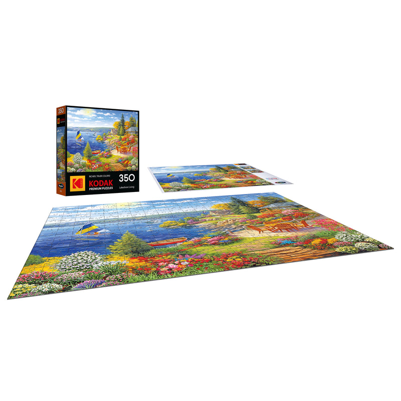 The ROSEART-KODAK PREMIUM "Lakeshore Living" 350-piece jigsaw puzzle showcases a vibrant lakeside scene with trees, flowers, a boat, and mountains; both the box and completed puzzle are included.