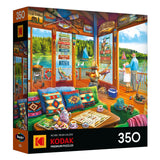 The ROSEART-KODAK PREMIUM-THE LAKE HOUSE 350 piece jigsaw puzzle features a vibrant lakeside cabin scene with people on a dock and cozy interior decor.