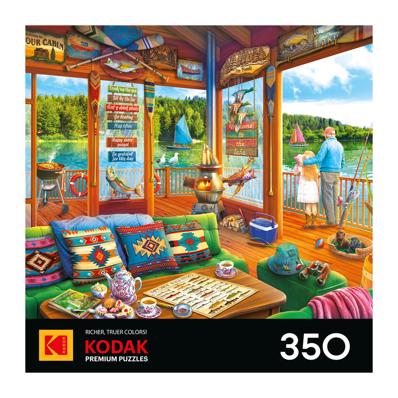 Colorful lake house cabin with ROSEART-KODAK PREMIUM-THE LAKE HOUSE-350 PIECE JIGSAW PUZZLE and tea on the table, overlooking a lake where a man and girl fish from the porch as sailboats pass by.