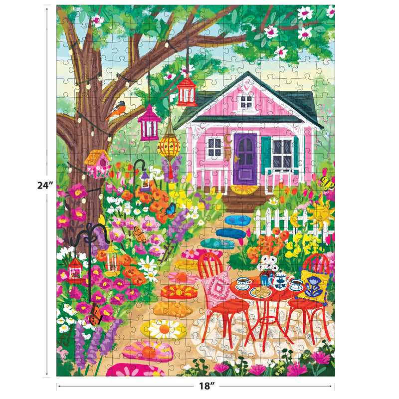The ROSEART-OLIVIA GIBBS-FLORAL GARDEN 300 piece jigsaw puzzle features a colorful scene with a pink house, flowers, lanterns, a tree, and patio furniture. Finished size is 24 x 18 inches.