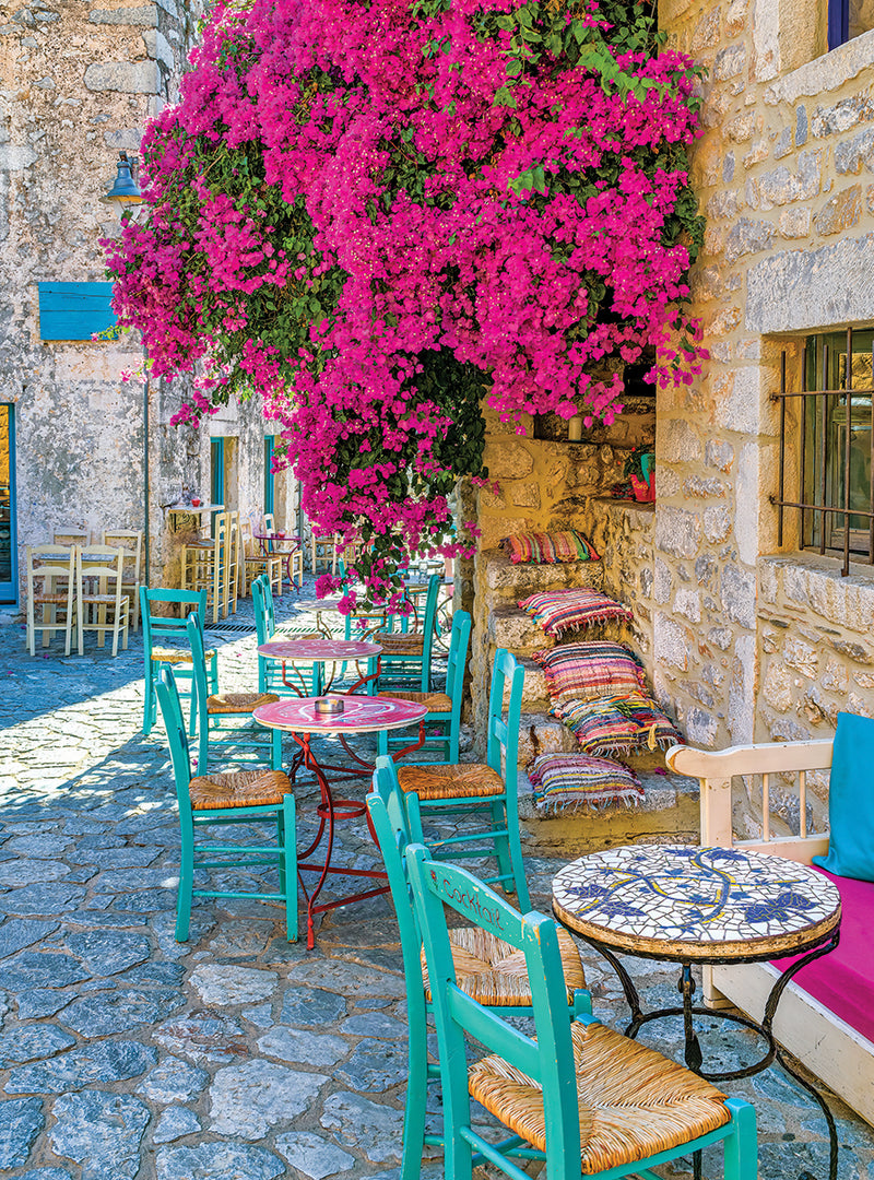 Outdoor cafe with vibrant pink bougainvillea, turquoise chairs, and stone-paved pathway. Tables have colorful patterns, and multicolored cushions are on a stone bench by the wall. The scene resembles the picturesque image captured in the RoseArt - Kodak Premium - Village Areopoli - 1000 Piece Jigsaw Puzzle.