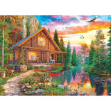 A picturesque log cabin with a stone pathway sits beside a tranquil river. A canoe is docked at the riverbank, surrounded by lush greenery and a vibrant sunset sky with birds flying, just like in the RoseArt - My Happy Place - Forest Cabin Home - 1000 Piece Jigsaw Puzzle.