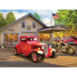 A classic red car and a motorcycle are parked in front of an old garage with vintage signage. Additional classic cars are seen parked next to the building. An American flag is flying above the garage, depicted in the RoseArt - American Nostalgia - Hot Rod Flames - 550 Piece Jigsaw Puzzle.