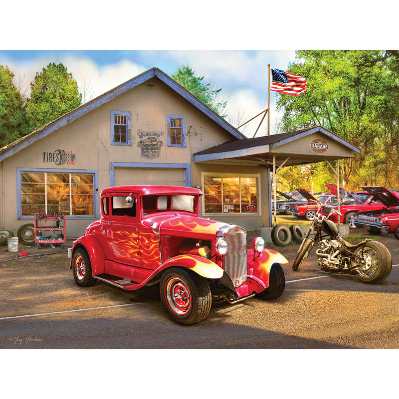 A classic red car and a motorcycle are parked in front of an old garage with vintage signage. Additional classic cars are seen parked next to the building. An American flag is flying above the garage, depicted in the RoseArt - American Nostalgia - Hot Rod Flames - 550 Piece Jigsaw Puzzle.