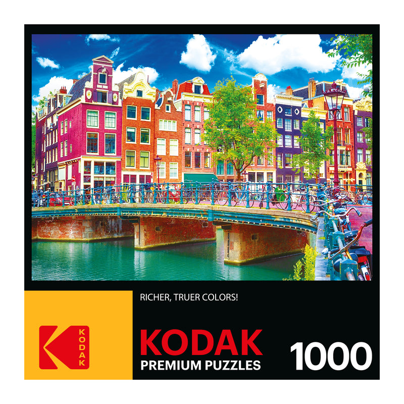 A colorful puzzle box labeled "RoseArt - Kodak Premium - Colorful Waterfront Buildings, Amsterdam - 1000 Piece Jigsaw Puzzle," featuring an image of a vibrant cityscape with multicolored buildings, a canal, and a bridge with bicycles. Text reads "Richer, truer colors!".