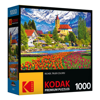 A box of RoseArt - Kodak Premium - Brienz Town and Flowers, Switzerland - 1000 Piece Jigsaw Puzzle with an image of a colorful landscape featuring bright tulips, houses, trees, and snow-capped mountains. The puzzle has 1000 pieces.