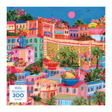 The ROSEART - MILLIE ILLUSTRATES - MARSEILLE 300-piece puzzle features a vibrant illustration of a Mediterranean-style town with colorful buildings, lush plants, and a bright pink sun—an ideal addition to any Millie Illustrates collection or as an engaging jigsaw for puzzle enthusiasts.