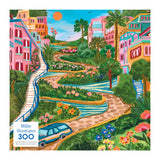 The ROSEART - MILLIE ILLUSTRATES 300-piece jigsaw puzzle depicts a vibrant Lombard Street scene in San Francisco, with lush vegetation, colorful buildings, and a car on the winding cobblestone path.