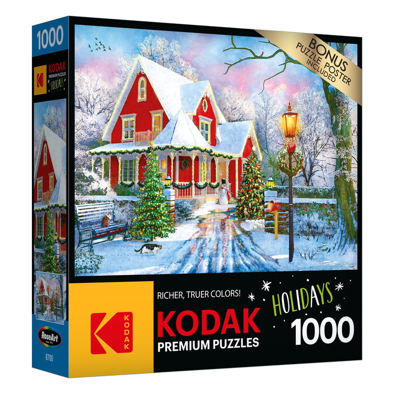 Box of a RoseArt - Kodak Premium - Christmas at Home - 1000 Piece Jigsaw Puzzle featuring a snowy holiday scene with a red house, decorated tree, lamppost, and a fence.