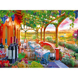 Outdoor patio with tables set for dining, surrounded by colorful flowers and grapevines. Bottles of wine, fruits, and a scenic vineyard landscape with hills and a distant path are visible in the RoseArt - Kodak Cork - Wine Country - 1000 Piece Jigsaw Puzzle.