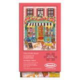 The ROSEART - VERTICAL VISIONS - The Story Bean is a 500 piece jigsaw puzzle showing a lively bookstore café scene with a bicycle, flowers, cats, and outdoor tables—perfect for relaxing afternoons.