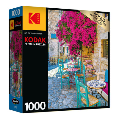A RoseArt - Kodak Premium - Village Areopoli - 1000 Piece Jigsaw Puzzle box showing a 1000-piece puzzle with an image of an outdoor café setting featuring colorful chairs, vibrant flowers, and stone buildings. Bonus puzzle poster included.
