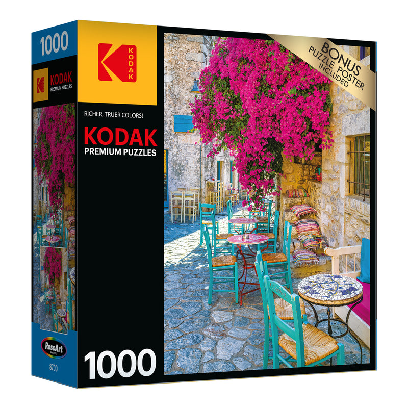 A RoseArt - Kodak Premium - Village Areopoli - 1000 Piece Jigsaw Puzzle box showing a 1000-piece puzzle with an image of an outdoor café setting featuring colorful chairs, vibrant flowers, and stone buildings. Bonus puzzle poster included.