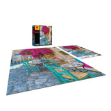 A partially completed jigsaw puzzle on a table depicts an outdoor café scene with teal chairs and pink flowers. The *RoseArt - Kodak Premium - Village Areopoli - 1000 Piece Jigsaw Puzzle* box and the puzzle image reference sheet are placed nearby.