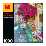 A 1000-piece RoseArt - Kodak Premium - Village Areopoli - 1000 Piece Jigsaw Puzzle box featuring an outdoor cafe with vibrant turquoise chairs, stone pavement, and blooming magenta flowers. Bonus puzzle poster included.