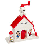 A Snoopy Snow Cone Maker featuring a red roof and decorated with cartoon characters. Snoopy sits atop the roof with a red handle, and a red cup and shovel are at the base.