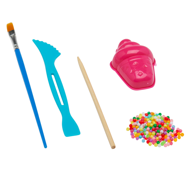 A set of tools and materials including a paintbrush, a blue plastic tool, a wooden stick, a pink shell-shaped mold, and a pile of colorful beads from the Cra-Z Crackle Clay.