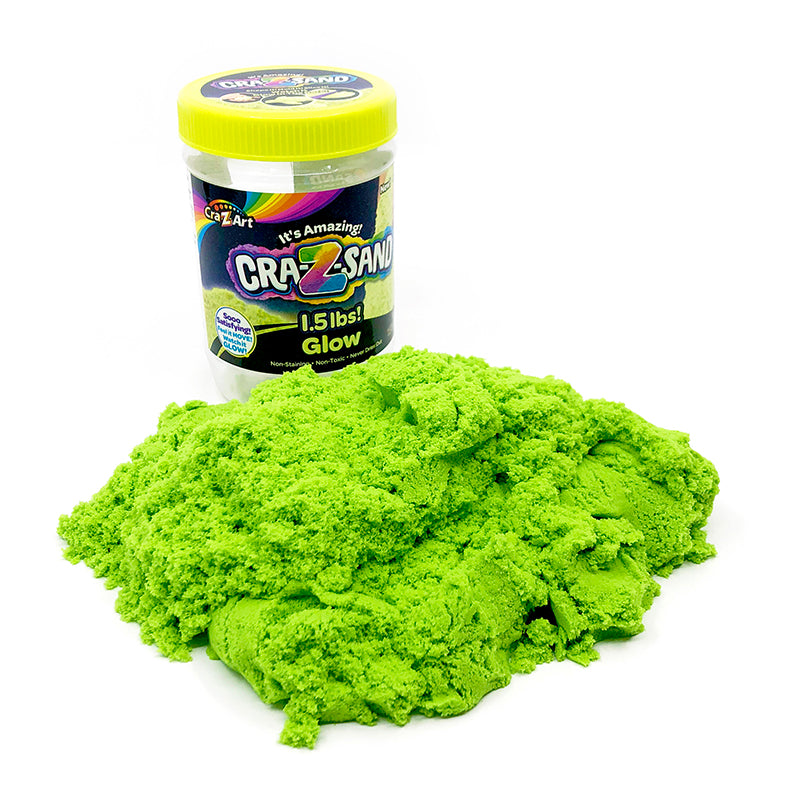 A container of Cra-Z Sand Glow sits behind a pile of green kinetic sand. The container label shows the weight of 1.5 lbs and the brand's logo.